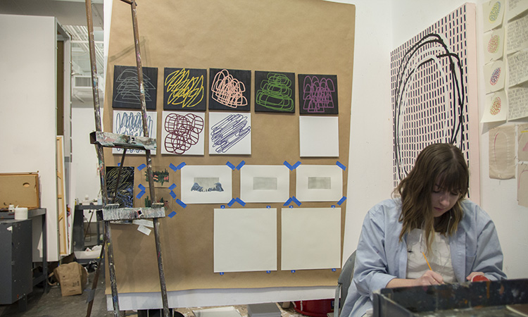 A student painting in front of a grid of small paintings