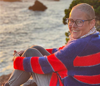 Silke Otto-Knapp sitting in front of the ocean wearing a red and blue striped sweater. Photo credit: Sharon Lockhart