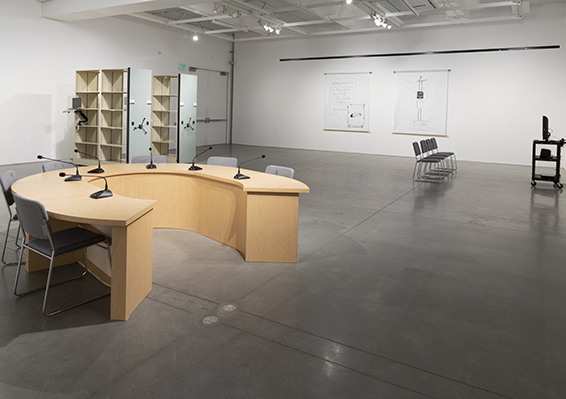 Installation view of graduate student work in the New Wight Gallery. A semi-circular desk is in the foreground with charts and bookshelves in the background.