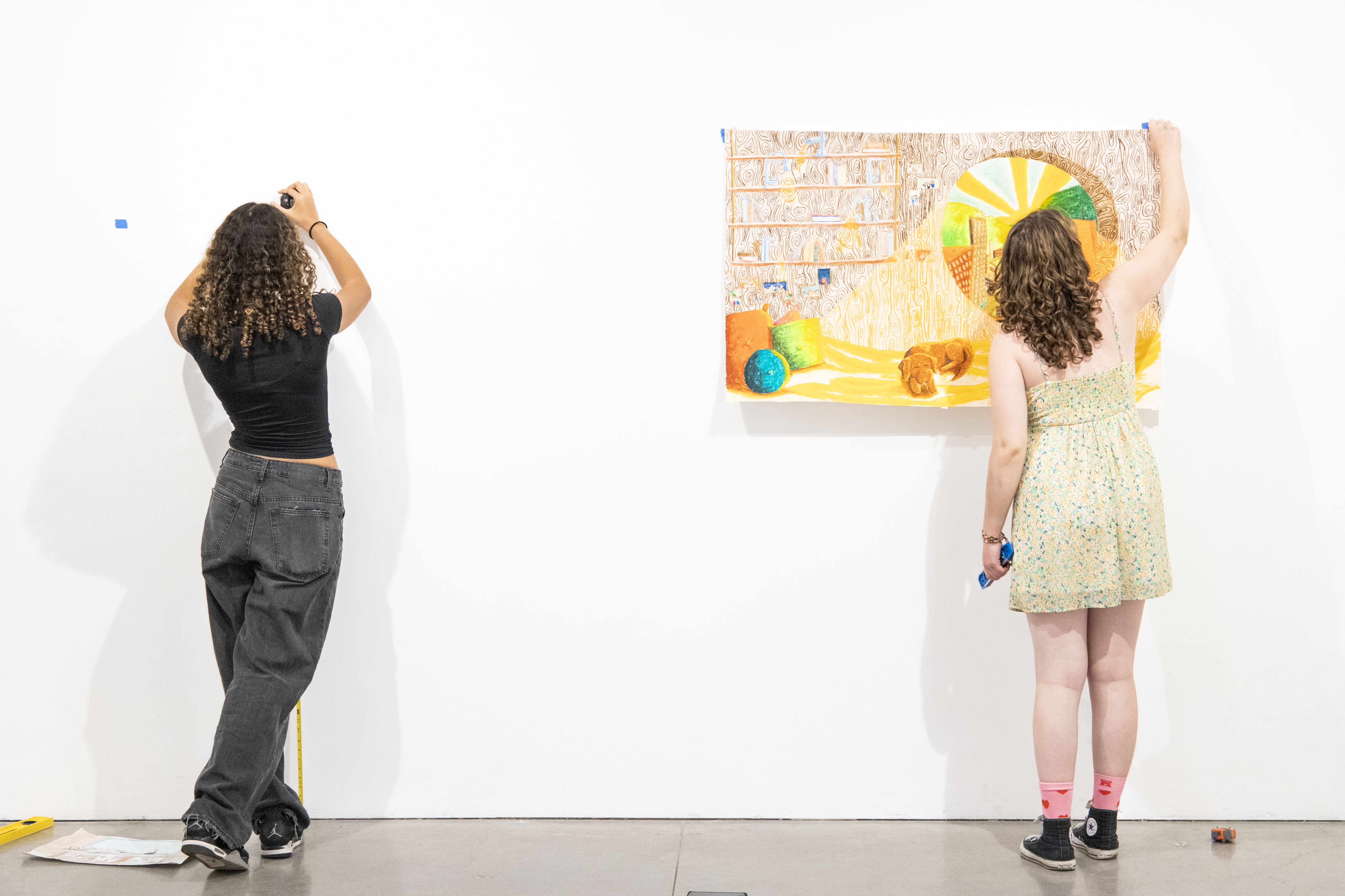 Two students adding tape to a blank, white wall to mount their artwork.